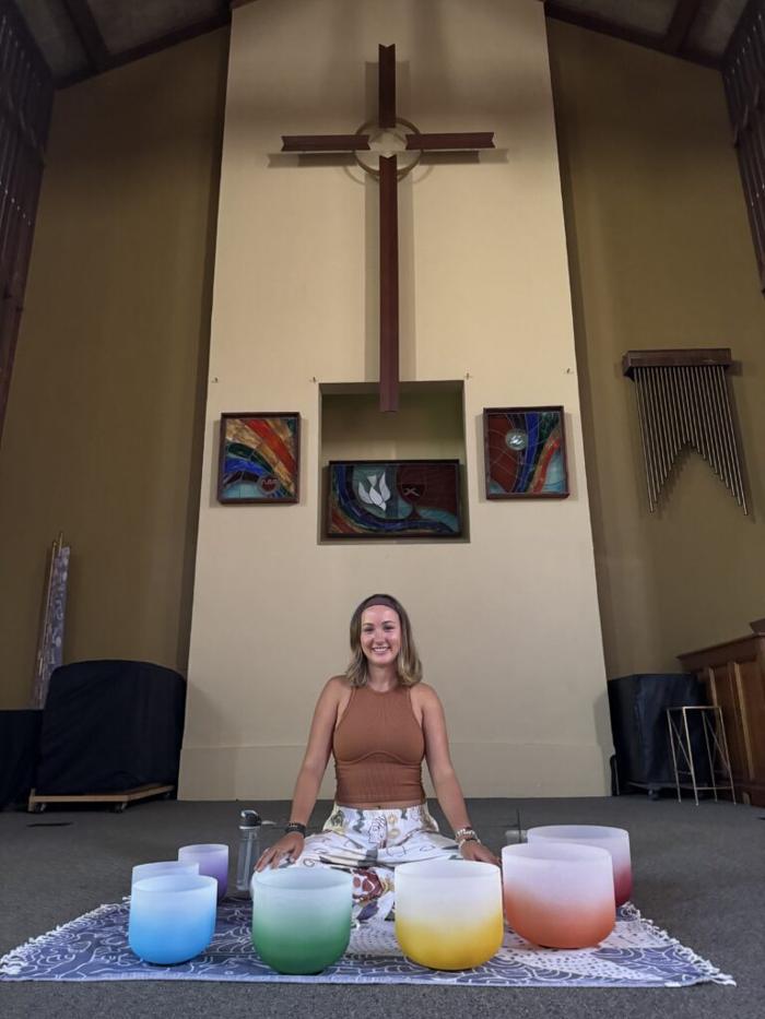 A young woman sitting cross-legged on the floor of the church sanctuary in front of a large wooden cross, surrounded by a circle of colorful singing crystal bowls.