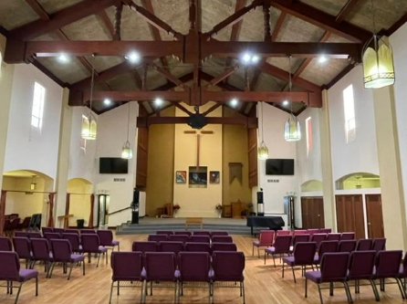 a big hall room with a small stage in front. the building has big white walls and a brown, open truss roof.